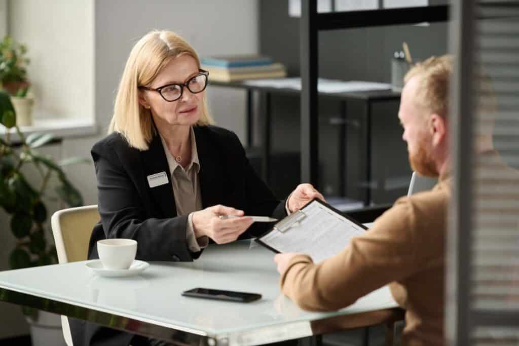 Eine Frau mit Brille und schwarzem Blazer sitzt an einem Tisch, hält ein Klemmbrett in der Hand und unterhält sich mit einem bärtigen Mann ihr gegenüber in Versicherungsberatung. Eine Tasse und ein Smartphone stehen auf dem Tisch, umgeben von üppigen Pflanzen im Hintergrund.