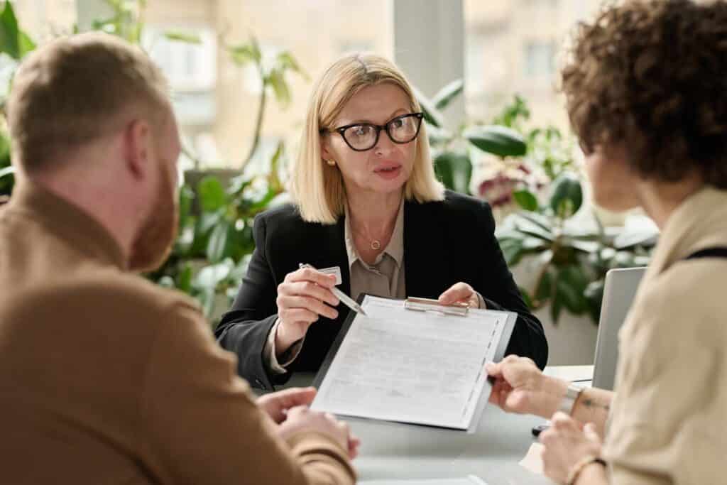 Eine Frau mit Brille hält ein Klemmbrett in der Hand und unterhält sich mit zwei Personen am anderen Tisch. Sie befinden sich in einem hellen, mit Pflanzen geschmückten Raum. Als unabhängiger Versicherungsmakler berät sie die Betroffenen kompetent und individuell.
