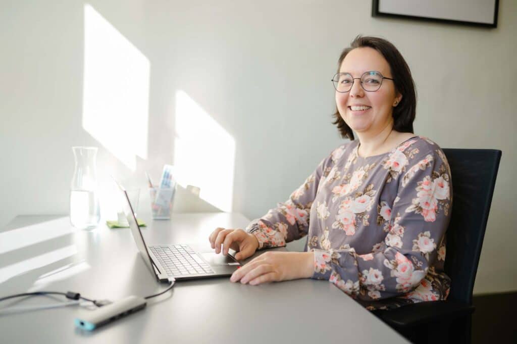 Eine lächelnde Person mit Brille und geblümter Bluse sitzt an einem Schreibtisch und tippt auf ihrem Laptop. Sonnenlicht fällt durch das Fenster und wirft Muster an die Wand. Daneben stehen eine Glaskaraffe und Schreibwaren, was darauf schließen lässt, dass die Person in der Online-Versicherungsberatung tätig ist und Kunden berät.