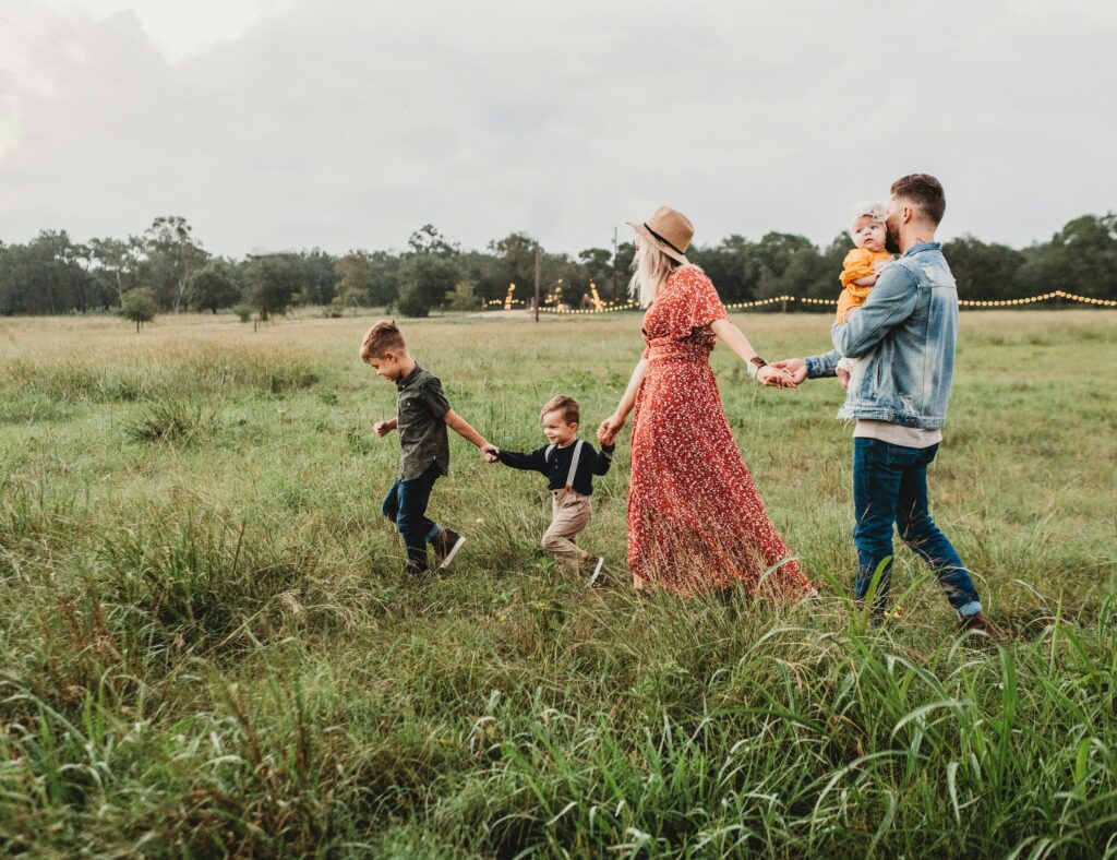 Eine fünfköpfige Familie geht Hand in Hand über eine Wiese und verkörpert damit die Bedeutung von „Familie absichern“. Die Mutter trägt ein rotes Kleid und einen roten Hut, der Vater hält ein Kleinkind, und zwei Jungen gehen voran. Im Hintergrund leuchten Bäume und Lichterketten.